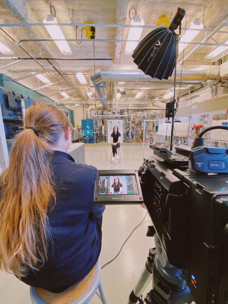 Interview with student sitting in manufacturing lab, while in focus on viewfinder, as Sara Sampey sits on the left of the professional camera.