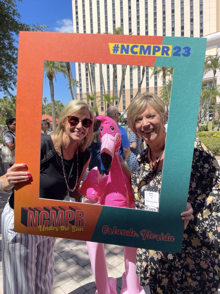 Mary and Diane pose with a pink flamingo inside of a NCMPR 2023 frame.