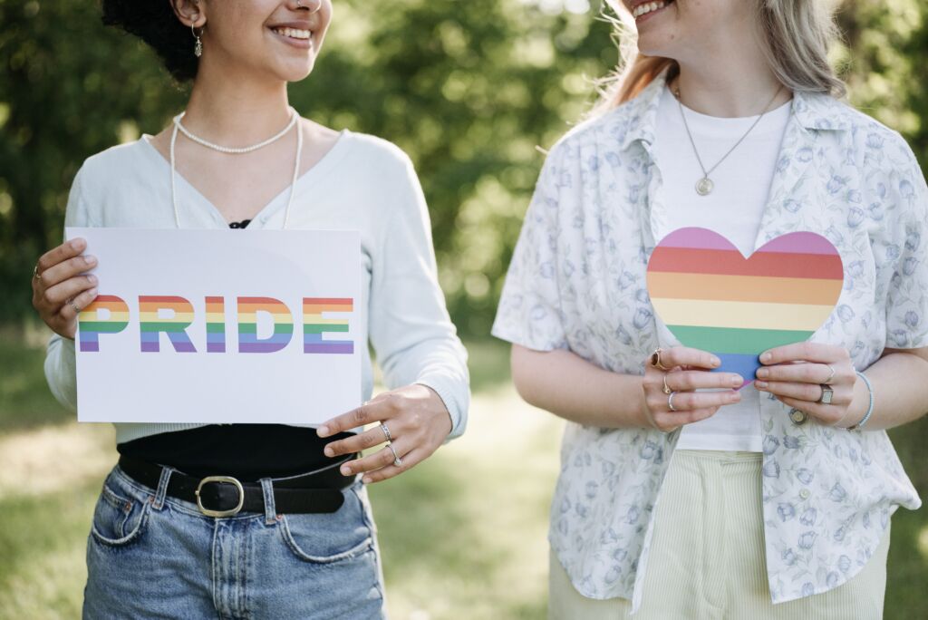 A lesbian couple holds up rainbow Pride signs. When we ask What's LGBTQ?, Lesbian is the first term. lgbtq meaning is important in higher ed right now and beyond!